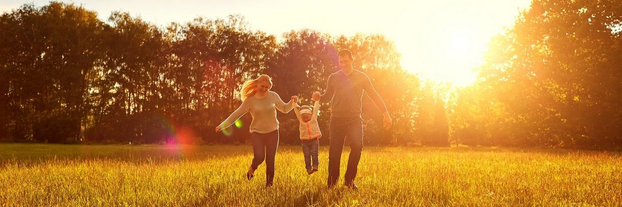 Familie läuft Hände haltend über ein Feld. Im Hintergrund geht die Sonne unter.