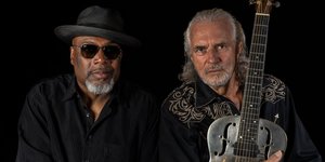 Hans Teessink und Big Daddy Wilson sitzen vor schwarzem Hintergrund und blicken ernst in die Kamera. Theessink hält eine silberne Gitarre.