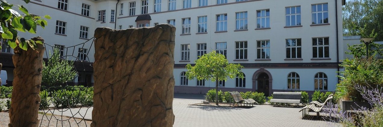 Historischer Schulhof mit Kletterelementen, Sitzbänken und einem hellen Schulgebäude im Hintergrund.