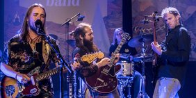 Drei Männer spielen auf einer Bühne Gitarre, im Hintergrund ein Schlagzeuger