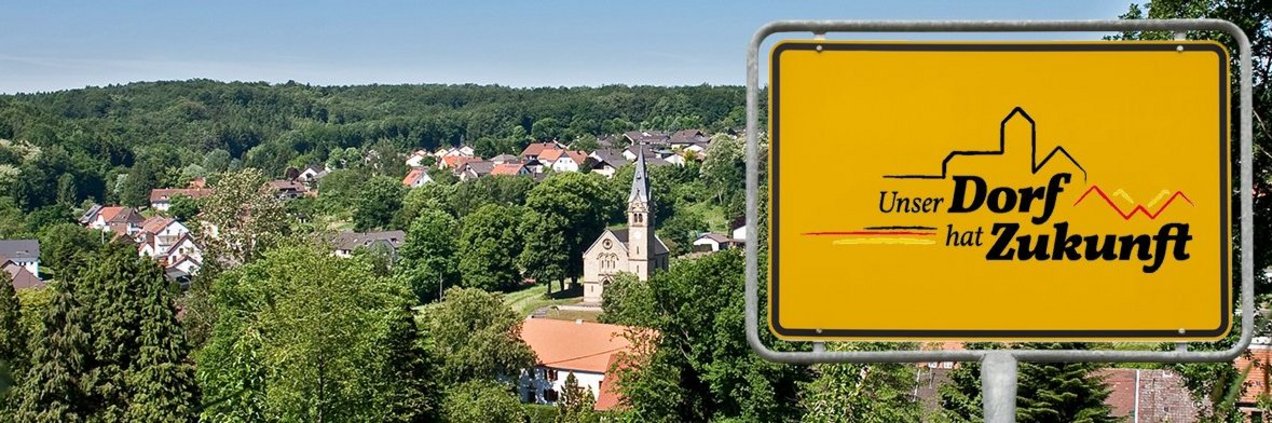 Schild "Unser Dorf hat Zukunft" Blick in ein Dorf im Tal. Im Vordergrund das Schild mit dem Aufdruck "Unser Dorf hat Zukunft".