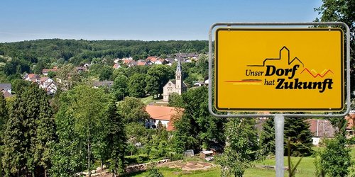 Blick in ein Dorf im Tal. Im Vordergrund das Schild mit dem Aufdruck "Unser Dorf hat Zukunft".