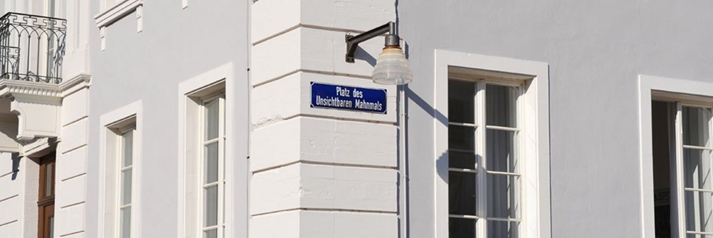 Schild am Schloss mit der Aufschrift Platz des Unsichtbaren Mahnmals