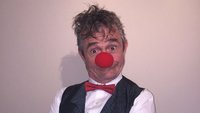 Ein Mann in Hemd, Sakko und roter Fliege trägt eine rote Clownsnase.