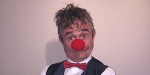 Ein Mann in Hemd, Sakko und roter Fliege trägt eine rote Clownsnase.
