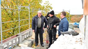 Der Erste Regionalverbandsbeigeordnete Norbert Moy und Bauamtsmitarbeiter Giuseppe Messina stehen auf dem Dach der Graf-Ludwig-Gemeinschaftsschule in Ludweiler und sprechen mit mehreren Männern einer Dachdeckerfirma, die dort gerade Sanierungsarbeiten vornimmt.