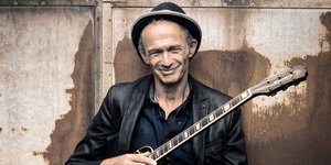 Mit der Gitarre auf dem Schoß lächelt Abi Wallenstein in die Kamera. Er trägt Hut und Lederjacke.