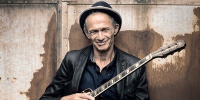 Mit der Gitarre auf dem Schoß lächelt Abi Wallenstein in die Kamera. Er trägt Hut und Lederjacke.