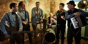 Sechs Männer in einem kleinen Raum beim Musizieren. An der Wand hinter ihnen hängt die Figur eines weißen Hais.
