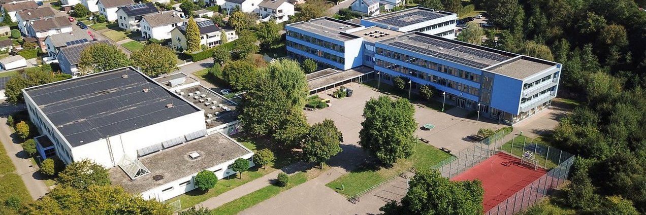 Luftaufnahme eines Schulgeländes in Heusweiler mit mehreren Gebäuden, Schulhof, Bäumen und rotem Sportplatz; im Hintergrund sind Wohnhäuser und die umliegende Landschaft zu sehen.