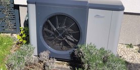 Blick auf eine Wärmepumpe an einer Hauswand leicht verdeckt von Rosmarinbüschen