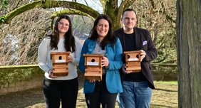 Regionalverbandsdirektorin Dr. Carolin Lehberger steht mit Elisa Haffner und Marc Mitzel von der Tourist Info Saarbrücker Schloss im Schlossgarten und alle halten je einen Nistkasten in den Händen. Im Hintergrund ist ein Nistkasten an einer Winterlinde zu sehen.