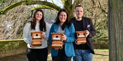 Regionalverbandsdirektorin Dr. Carolin Lehberger steht mit Elisa Haffner und Marc Mitzel von der Tourist Info Saarbrücker Schloss im Schlossgarten und alle halten je einen Nistkasten in den Händen. Im Hintergrund ist ein Nistkasten an einer Winterlinde zu sehen.